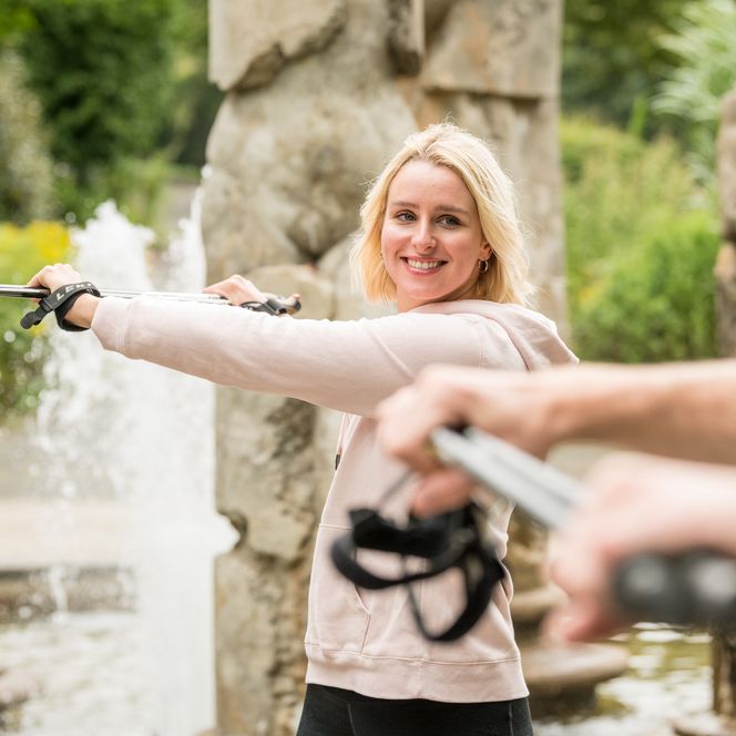 <p>Frau im Freien lächelt beim Training mit Stöcken vor einem Brunnen in entspannter Umgebung.</p>