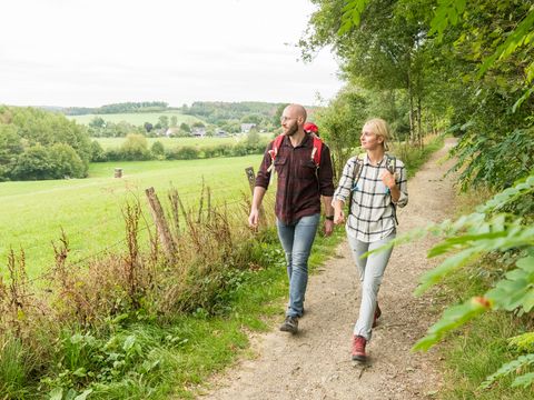 Ein junges Paar wandern über einen Waldweg. Am rechten Wegesrand stehen Bäume und Sträucher, am linken ist eine Wiese mit Fernblick über grüne Hügel und Wälder.