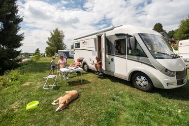 Wohnmobilparken auf grasbewachsener Fläche, Familie genießt Mahlzeit im Freien neben schlafendem Hund.