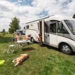 Wohnmobilparken auf grasbewachsener Fläche, Familie genießt Mahlzeit im Freien neben schlafendem Hund.