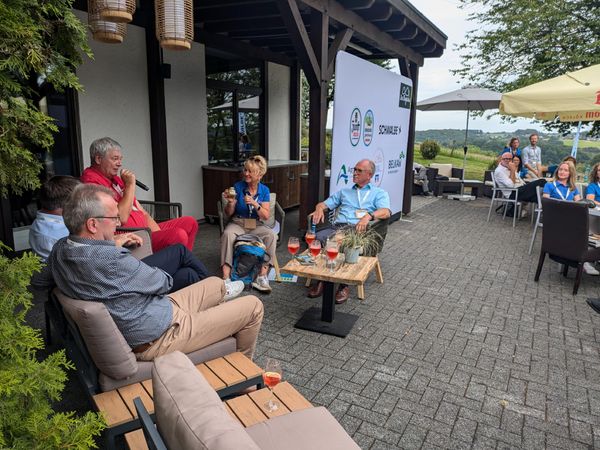 Fünf Teilnehmer einer Gesprächsrunde sitzen um einen Tisch gruppiert und diskutieren. Im Hintergrund sind Teilnehmer der Veranstaltung zu sehen und der Blick in die Landschaft.