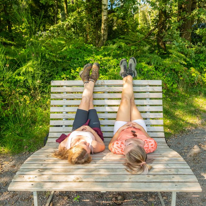 Zwei Personen liegen entspannt auf einer Holzbank im Wald, die Beine hochgelegt.