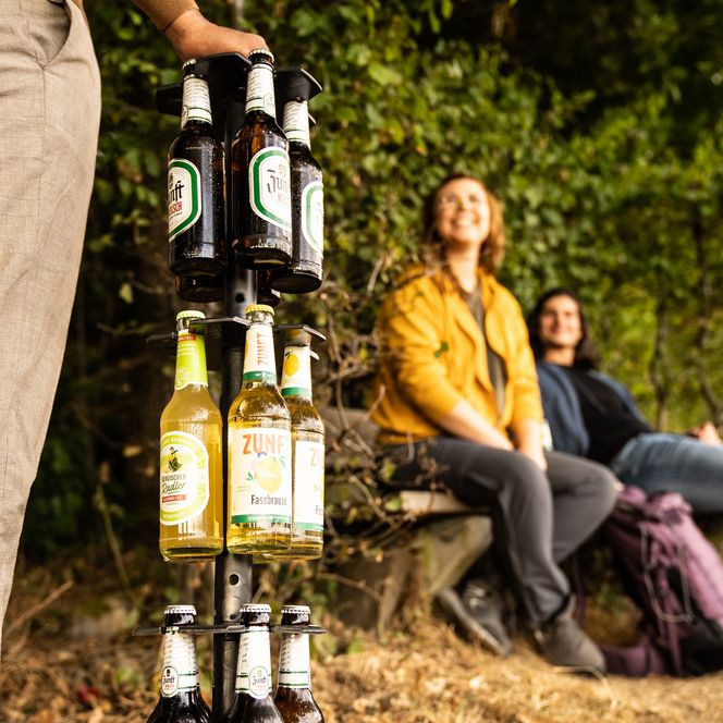 <p>Person hält Bier- und Limonadenflaschen im Freien, zwei Personen im Hintergrund auf einer Bank sitzend.</p>
