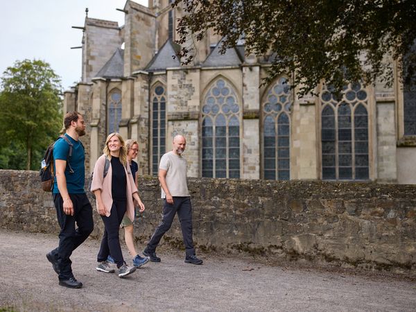 Zwei Frauen und zwei Männer wandern vor einer halbhohen Mauer. Im Hintergrund ist der Altenberger Dom zu sehen.