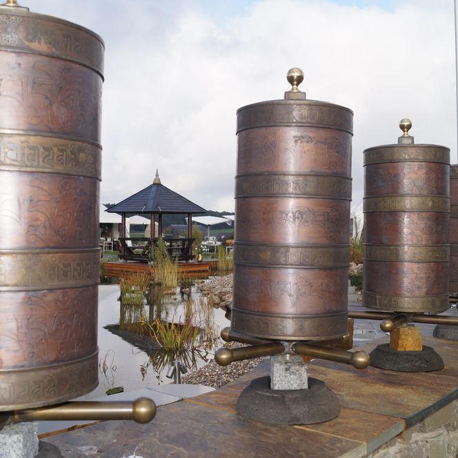 <img alt="Reihe von buddhistischen Gebetsmühlen aus Kupfer mit Pagode im Hintergrund" />