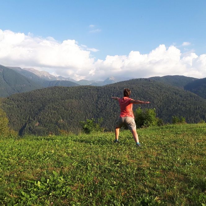 Eine Frau macht eine Yogaübung. Im Hintergrund ist weite, grüne Landschaft zu sehen.
