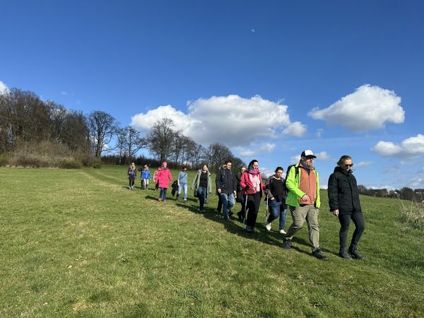 Eine Gruppe von Wandernden geht bei sonnigem Wetter über eine weite Wiese. Im Hintergrund stehen kahle Bäume und am Himmel sind Wolken und eine Mondsichel zu sehen.