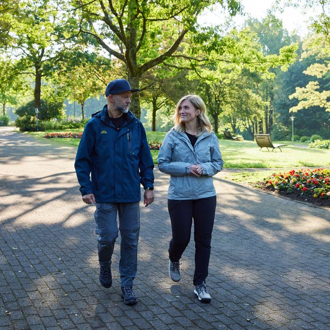 Zwei Menschen spazieren auf einem gepflasterten Weg in einem grünen Park bei sonnigem Wetter.