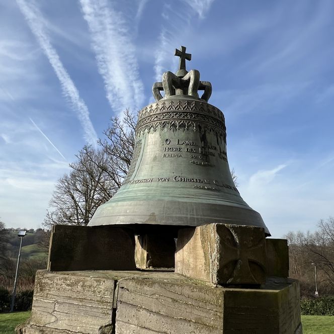 Die Gedenk-Glocke in Lohmar, montiert auf einem Holzsockel, vor blauem Himmel und Bäumen.