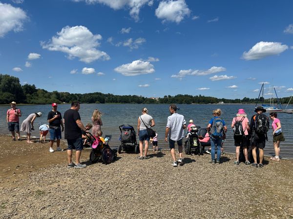 Eine Wandergruppe mit Menschen mit und ohne Rollstuhl steht am Ufer einer Talsperre. 