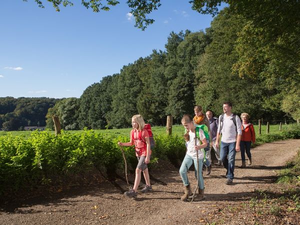 Eine Wandergruppe mit vier Erwachsenen und zwei Mädchen läuft über einen Wanderweg am Waldrand. Die Kinder laufen vorweg und haben Wanderstöcke in der Hand.