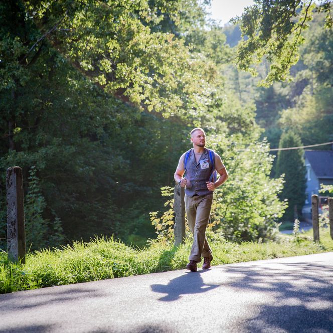Ein Mann geht bei Sonnenschein auf einer ländlichen Straße, umgeben von grüner Natur.