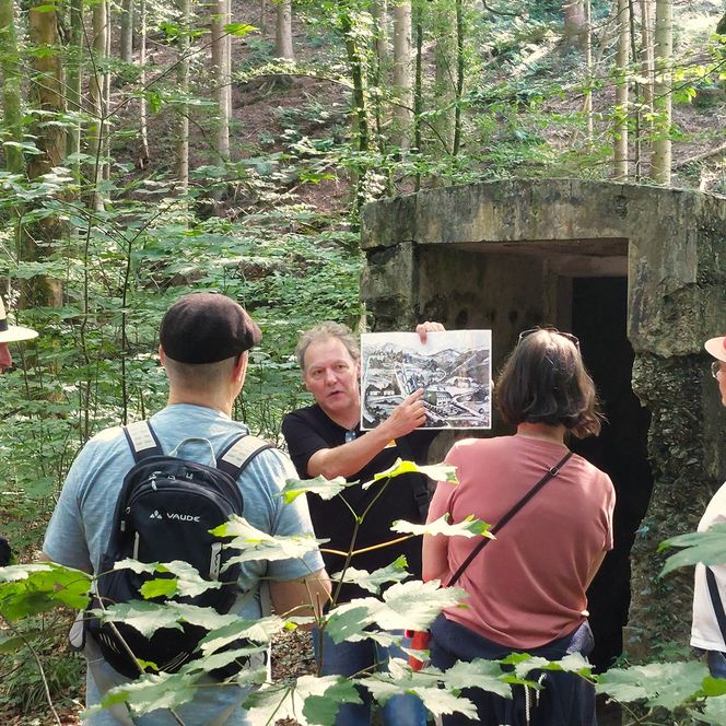 Eine Gruppe von vier Wandernden steht vor einem Gästeführer, der ein Bild in der Hand hält und darauf etwas erklärt.