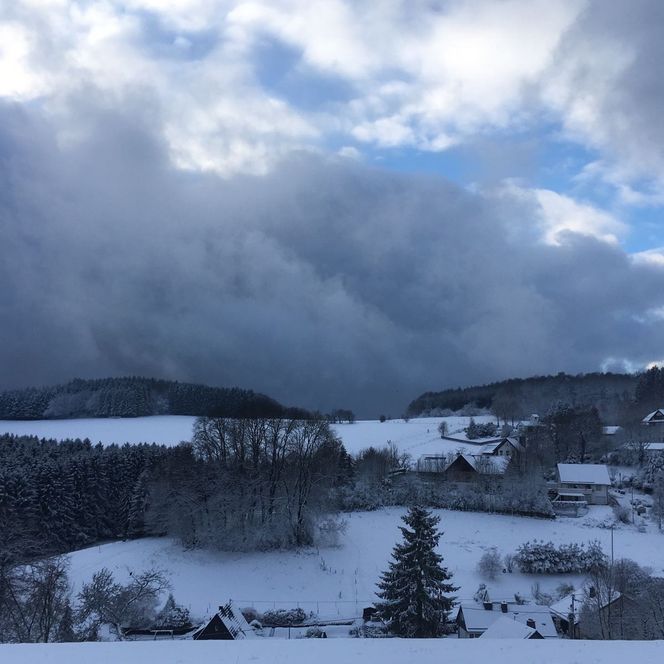 Schneebedeckte hügelige Landschaft mit verstreuten Häusern unter einem dramatischen, bewölkten Himmel.