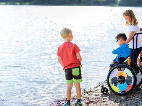 Vier Kinder stehen am Ufer einer Talsperre und schauen aufs Wasser. Ein Junge sitzt in einem Rollstuhl bei ihnen.