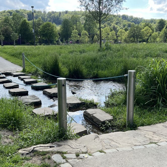 <p>Stufen aus Naturstein überqueren einen klaren Bach inmitten einer grünen, bewaldeten Landschaft.</p>