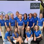 Die Mitarbeiterinnen von Das Bergischen stehen in blauen Poloshirts vor einer Leinwand.