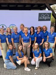 Die Mitarbeiterinnen von Das Bergischen stehen in blauen Poloshirts vor einer Leinwand.