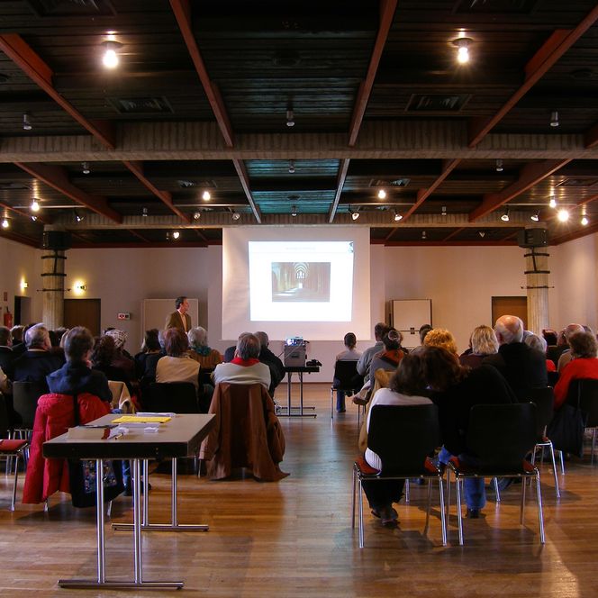 Ein Vortrag mit älteren Menschen in einem Saal; ein Redner steht vor einer Leinwand mit Projektion.