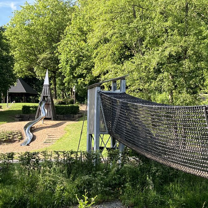 Eine Spielplatzlandschaft mit einer Rutsche aus Holz und einer Hängebrücke, umgeben von grünen Bäumen.