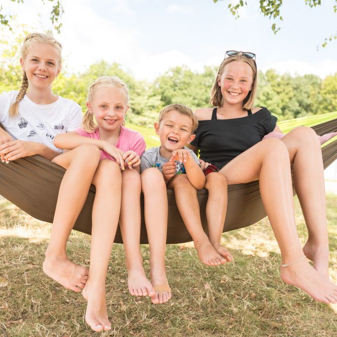 Familie sitzt lachend auf einer Hängematte im Freien, umgeben von sonnigem Wetter und grüner Natur.