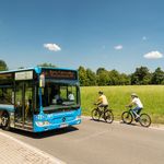 <p>Ein blauer RVK-Bus fährt an zwei Radfahrern entlang einer ländlichen Straße bei Sonnenschein.</p>