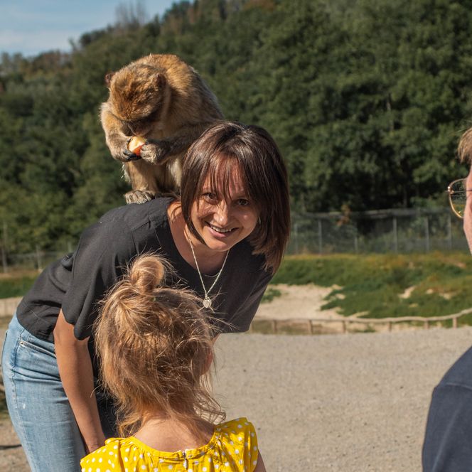 <p>Eine Frau lächelt, während ein Affe auf ihrer Schulter sitzt, umgeben von lachenden Menschen im Freien.</p>