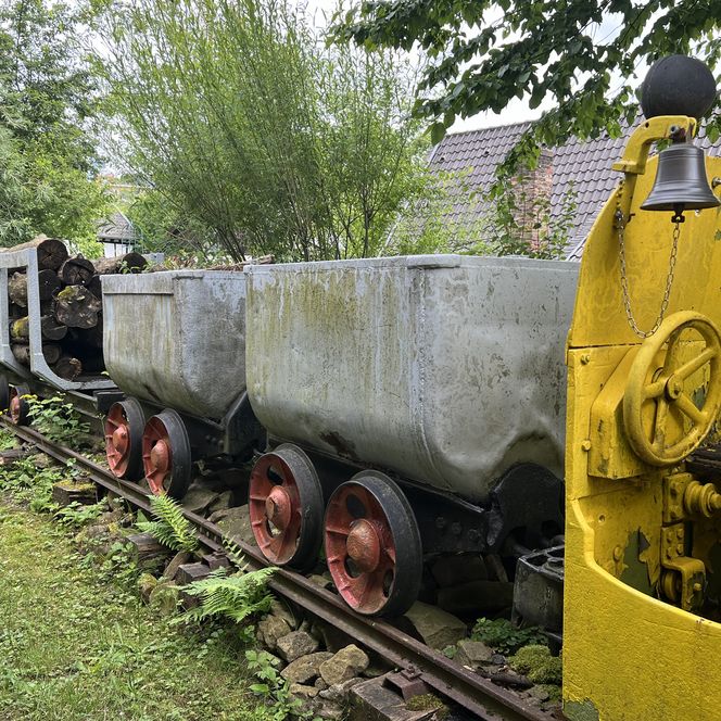 Die alten Bergwerkswagen auf Schienen sind von Bäumen umgeben, eine gelbe Lok steht daneben.