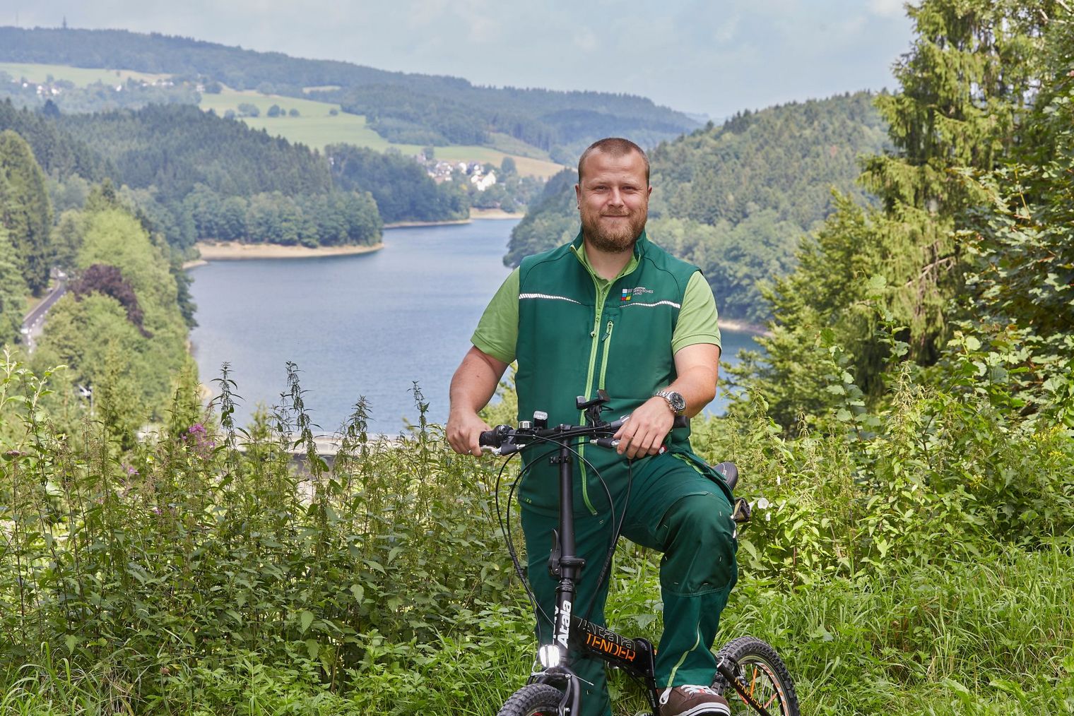 Foto des Wegepaten Andreas Kurze. Er sitzt auf einem Fahrrad auf einer Wiese. Hinter ihm ist eine Talsperre zu sehen.