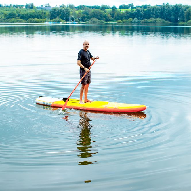 Mann auf gelbem SUP-Board paddelt auf ruhigem See, umgeben von grüner Landschaft und klarem Himmel.
