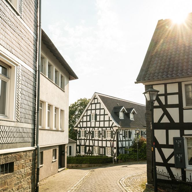 <p>Fachwerkhäuser in Ruppichteroth im Sonnenlicht, enge Kopfsteinpflasterstraße zwischen den Gebäuden.</p>