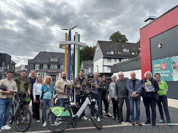 Gruppe von Menschen mit Fahrrädern und Broschüren vor einer farbenfrohen Skulptur und einem REWE-Markt