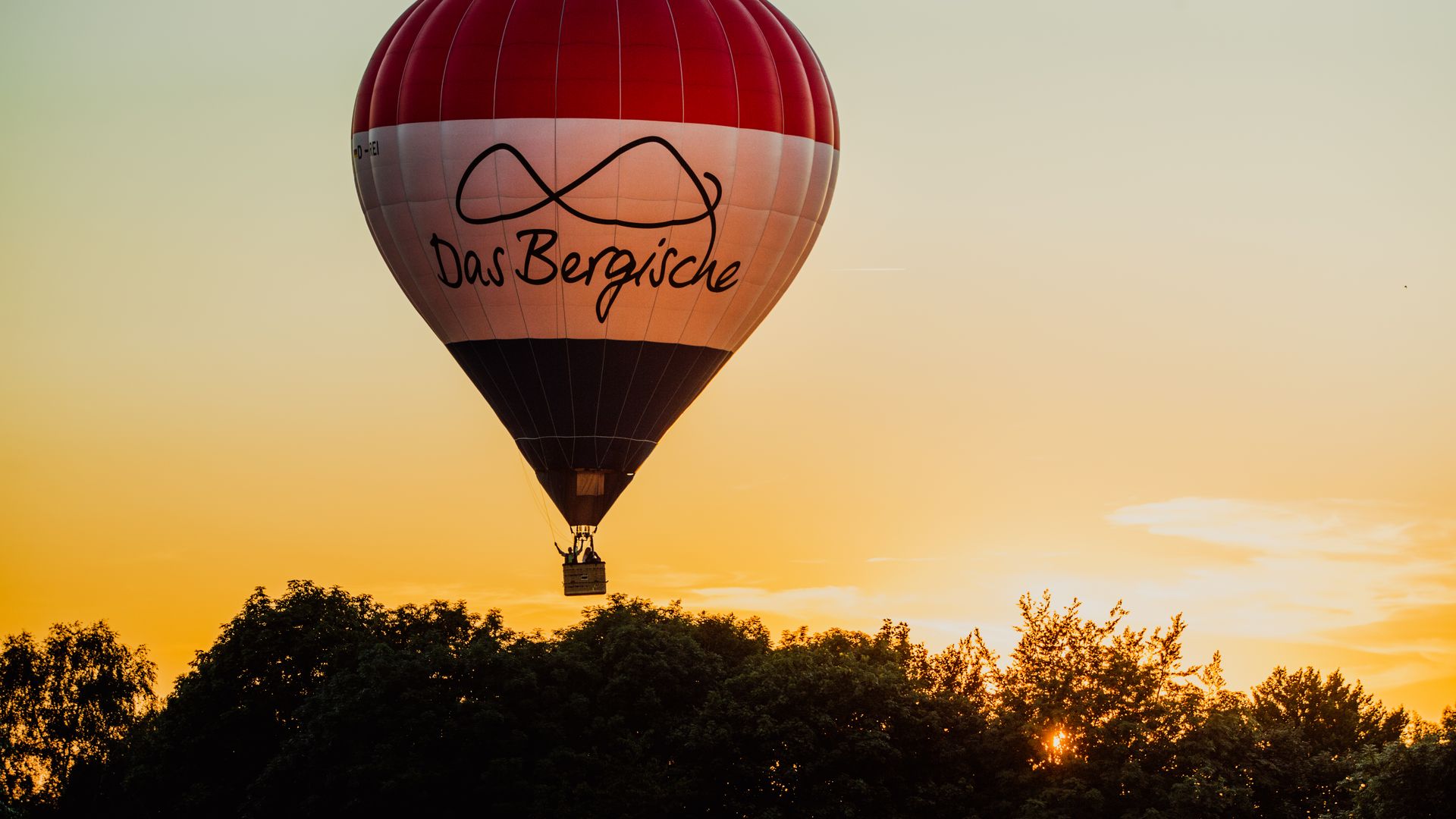 Ein Heißluftballon mit einem rot-weiß-blauen Ballon fährt über den Baumwipfeln im Sonnenuntergang.