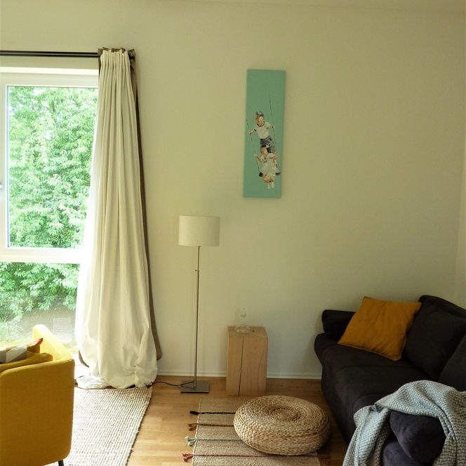 Helles Wohnzimmer mit gelbem Sessel, grauem Sofa, hellen Vorhängen und dekorativem Teppich.