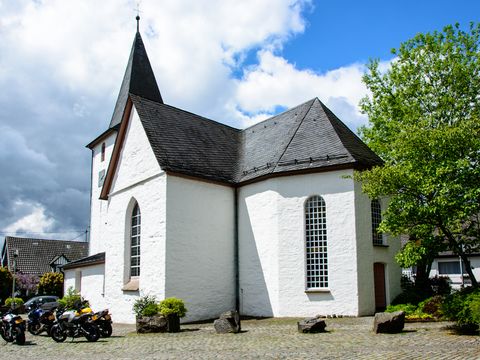 Eine kleine weiße Kirche mit einem dunklen Dach steht auf einem Platz mit einem Baum an der Seite. 