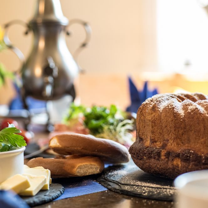 Ein gedeckter Tisch mit Gugelhupf, Brot, Butter und einer Kanne im Hintergrund.