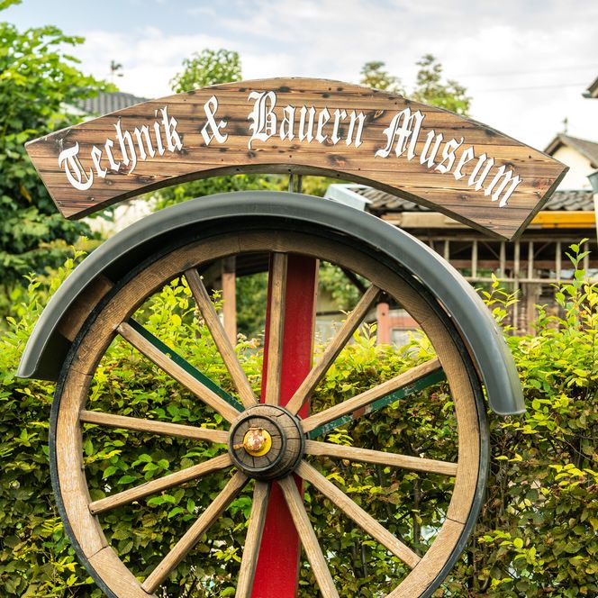 Technik & Bauern Museum Schild mit großem Holzwagenrad in grüner Gartenumgebung im Freien.