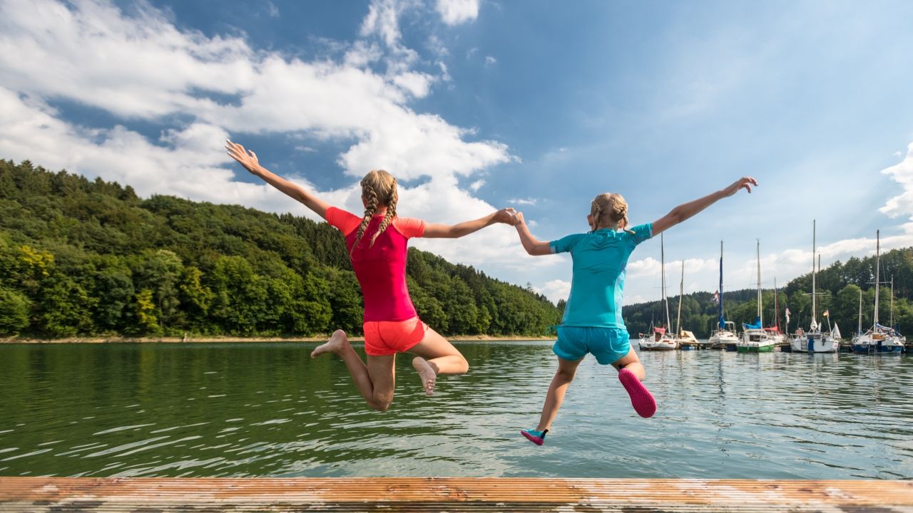 Zwei Mädchen springen vom Badesteg, von hinten zu sehen, Arme erhoben