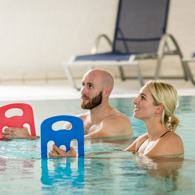 Zwei Personen in einem Schwimmbad, halten Schwimmbretter, Liegestühle im Hintergrund.