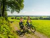 Zwei Radfahrer genießen eine Fahrt auf einem Waldweg mit weitläufigem Blick auf grüne Landschaften.