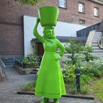 Grüne Statue einer Frau mit Eimer auf dem Kopf vor einem Backsteingebäude.