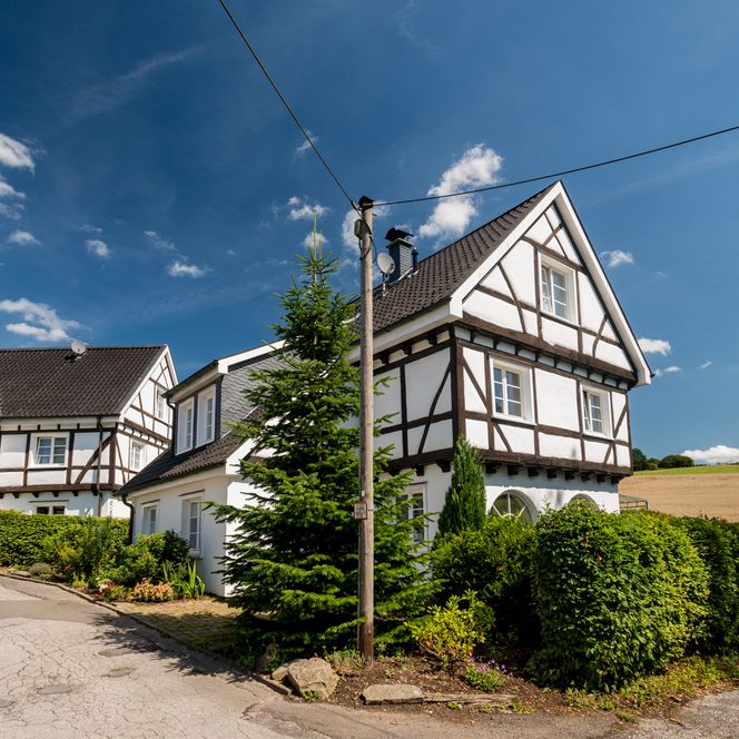 Fachwerkhäuser in Burscheid vor blauem Himmel, umgeben von grünen Sträuchern und Bäumen.