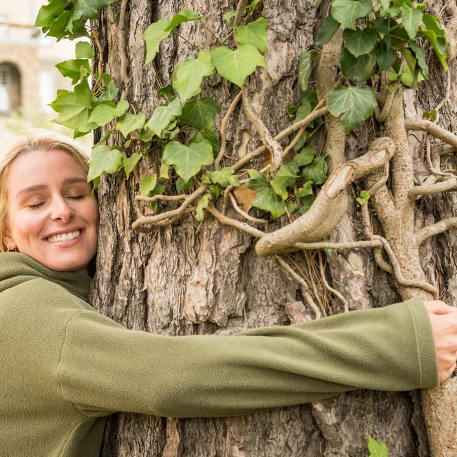 Eine lächelnde Frau umarmt einen großen Baum, überzogen von Kletterpflanzen, in einem Park.