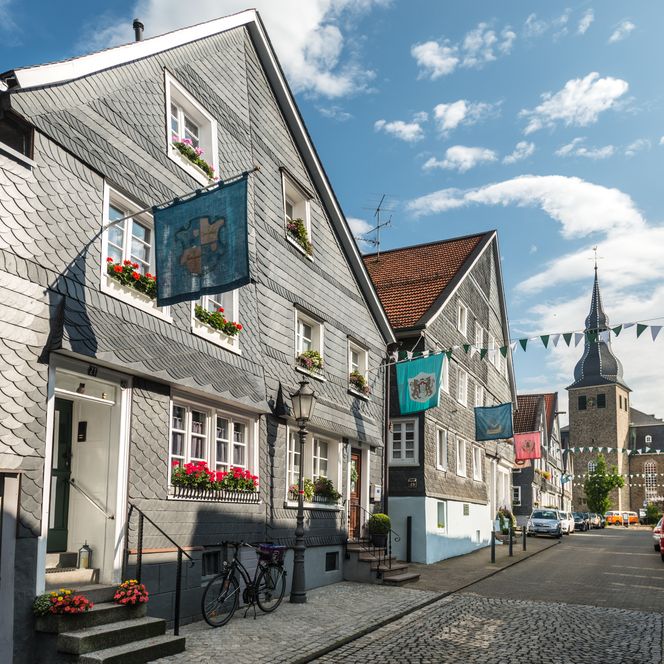Fachwerkhäuser mit bunten Fahnen in Hückeswagen, Kopfsteinpflasterstraße und Kirchturm im Hintergrund.