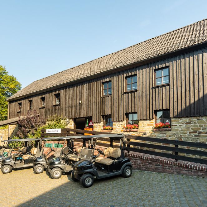 Golfcarts stehen vor einer rustikalen Holzhütte mit blühenden Blumen auf einem Pflastersteinplatz.