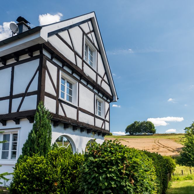 Fachwerkhaus mit weißer Fassade, umgeben von grünen Hecken und Feldern unter blauem Himmel.