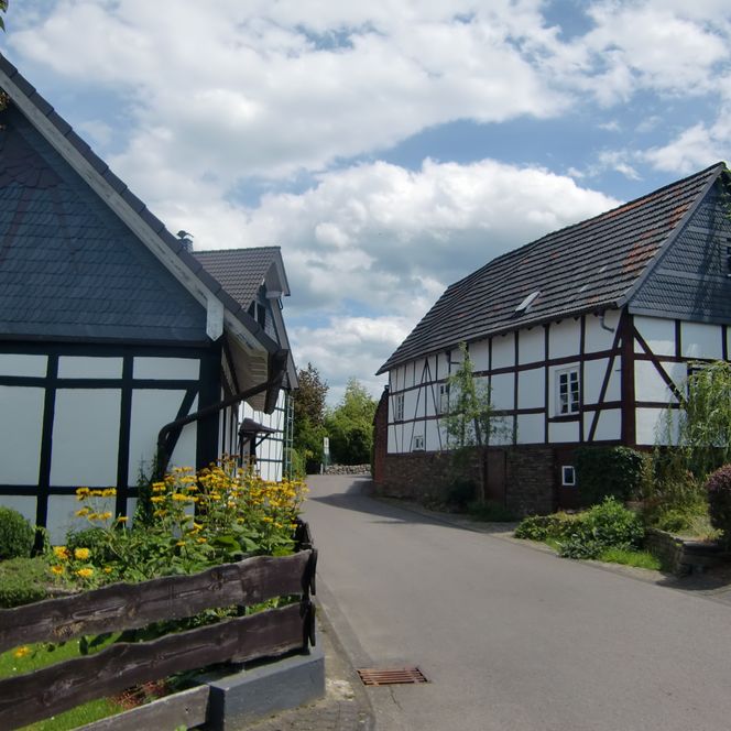 Fachwerkhäuser in einer ruhigen Straße in Wiehl, umgeben von Blumen und Bäumen, unter bewölktem Himmel.