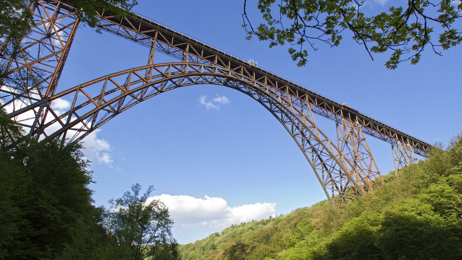 Blick vom Boden aus nach oben zu einer Eisenbahnbrücke über ein Tal. Im Hintergrund sind Bäume und blauer Himmel mit einigen Wolken zu sehen. 