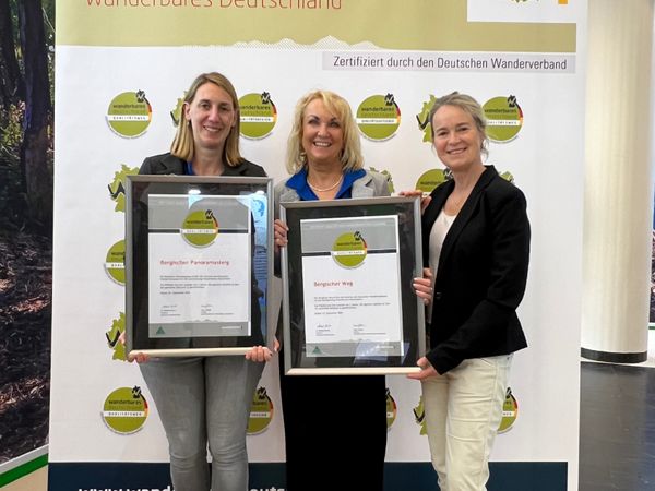 Drei Frauen stehen vor einer Leinwand und halten zwei Zertifikate in Bilderrahmen hoch.