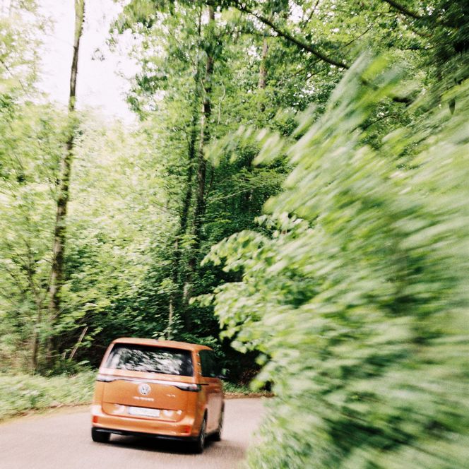 Ein orangefarbener Van fährt über eine Straße am Waldrand.